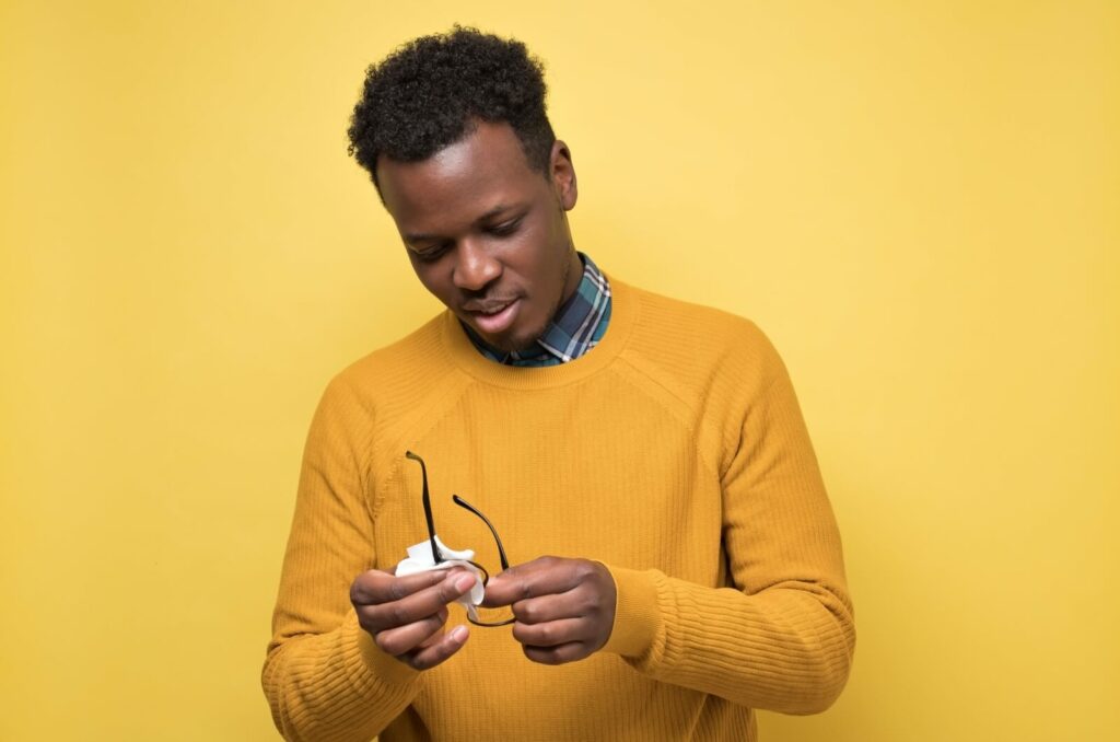 A person against a yellow background cleans their eyeglasses with a microfiber cloth.