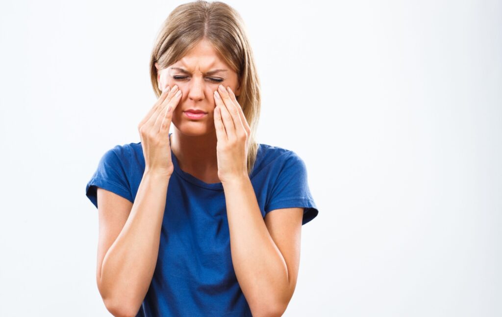 A person wearing a blue shirt presses their face with their hands while struggling to open their tired and sore eyes.