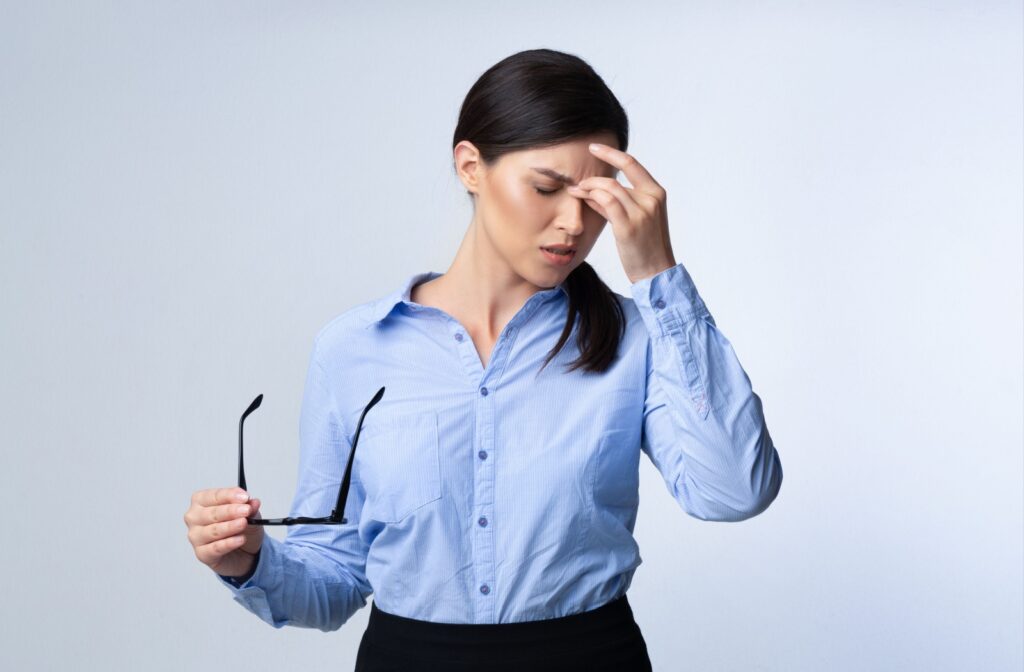 Woman in a blue shirt holding eyeglasses and massaging the bridge of her nose, appearing to have a headache, illustrating how vision problems like eye strain can lead to discomfort.