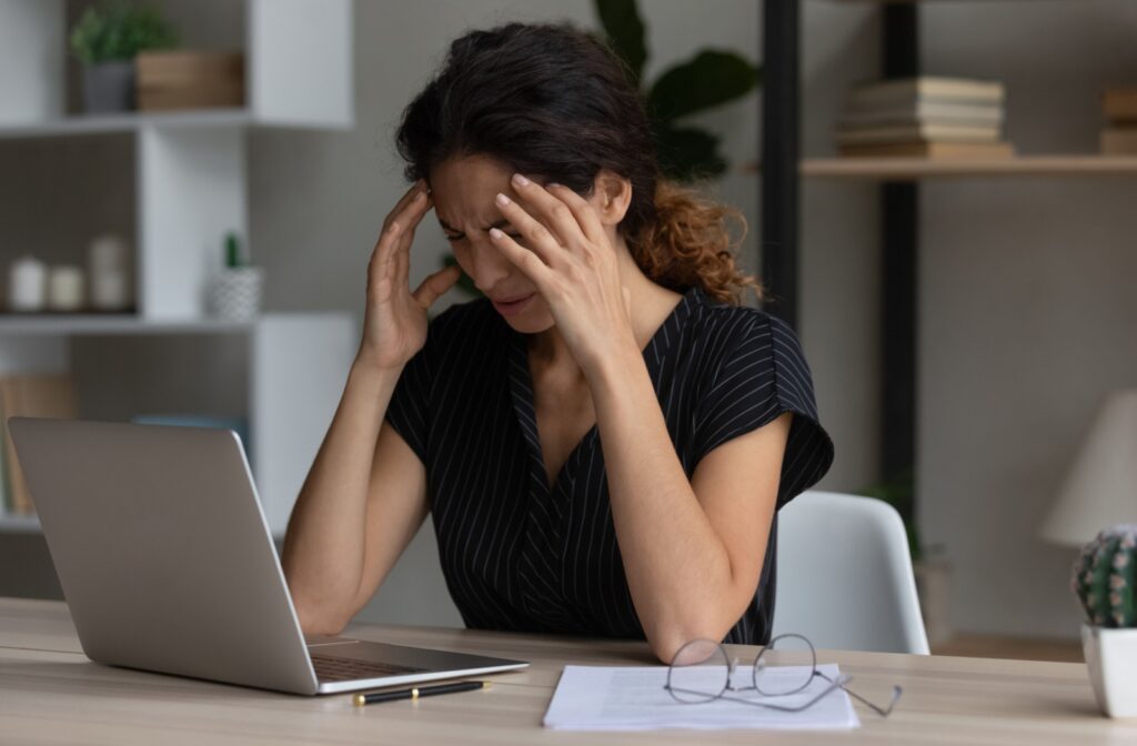 Woman sitting at a desk with a laptop, holding her temples in discomfort, suggesting a headache from prolonged screen use.