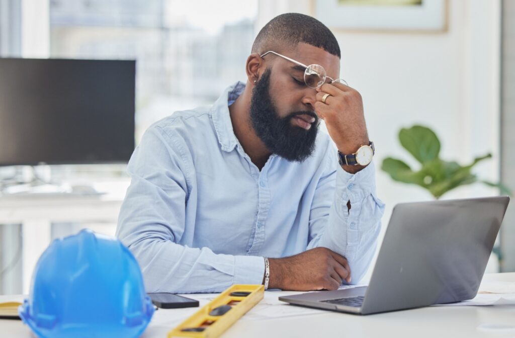 A man sitting at a desk with a computer screen in front of him, holding his glasses and rubbing his eyes, experience digital eye strain